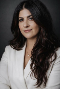 Woman with long dark wavy hair wearing a white blazer, posed against a dark background, looking at the camera with a neutral expression.